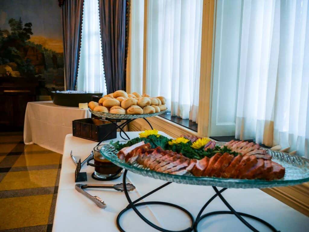 A buffet table by Baton Rouge Caterers features a platter of sliced meat garnished with parsley and yellow flowers in the foreground. Behind it, a plate of bread rolls is displayed. The table is placed near a window with sheer white curtains and brown drapes.