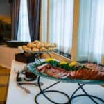 A buffet table by Baton Rouge Caterers features a platter of sliced meat garnished with parsley and yellow flowers in the foreground. Behind it, a plate of bread rolls is displayed. The table is placed near a window with sheer white curtains and brown drapes.