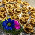 A platter of mini tostadas topped with seasoned ground meat and melted cheese is displayed on a silver tray. Garnished with a green leaf and three colorful flowers, this delightful appetizer showcases the artistry of 360 Catering Company. The vibrant presentation rests elegantly on a granite countertop.