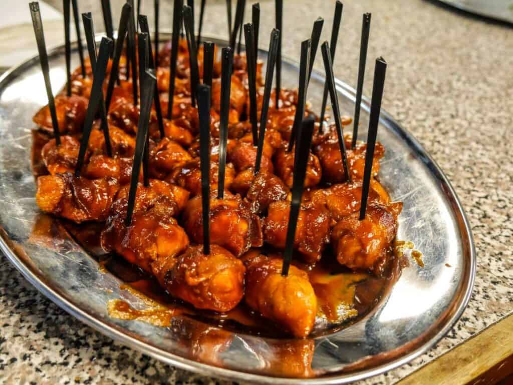 A metal serving tray with bite-sized pieces of food skewered on black plastic picks, glazed with a reddish barbecue or sweet-and-sour sauce, prepared by Baton Rouge Caterers. The tray is set on a speckled countertop surface, perfect for any event venue near Baton Rouge.