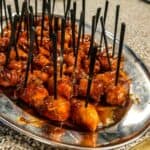 A metal serving tray with bite-sized pieces of food skewered on black plastic picks, glazed with a reddish barbecue or sweet-and-sour sauce, prepared by Baton Rouge Caterers. The tray is set on a speckled countertop surface, perfect for any event venue near Baton Rouge.