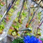 Small, clear plastic cups are filled with a diced mix of avocado and another ingredient topped with green onions, each containing a small spoon. They are arranged in rows on a tray by Baton Rouge Caterers, with a vibrant blue flower and a piece of green kale for decoration in the foreground.