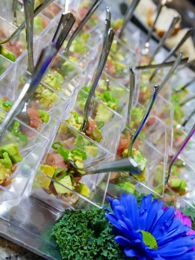 Small, clear plastic cups are filled with a diced mix of avocado and another ingredient topped with green onions, each containing a small spoon. They are arranged in rows on a tray by Baton Rouge Caterers, with a vibrant blue flower and a piece of green kale for decoration in the foreground.