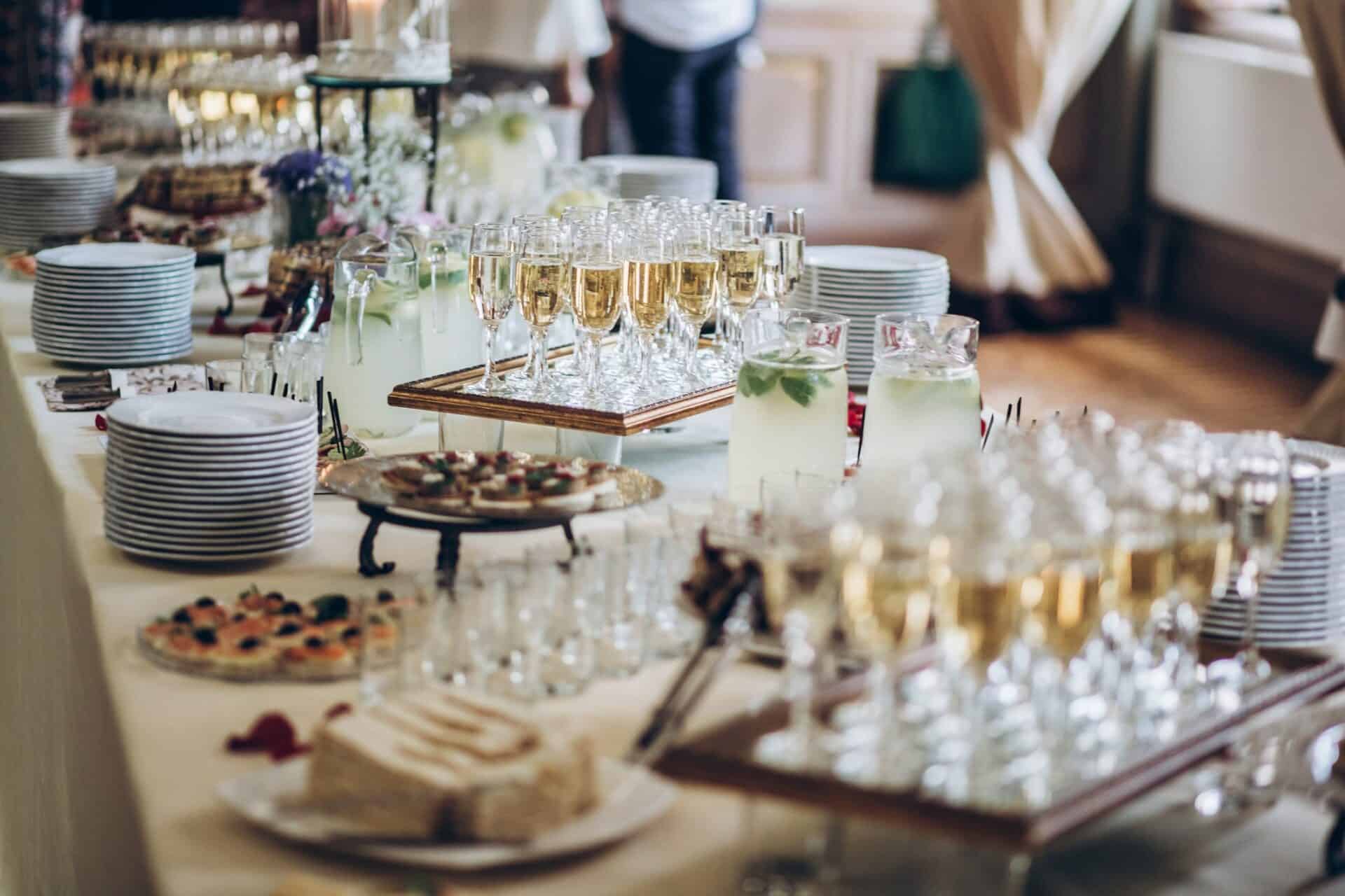 A long table is set with numerous champagne flutes filled with champagne, plates of hors d'oeuvres, and several pitchers of lemonade. Stacked plates and cutlery are also present, likely for guests to serve themselves at a Baton Rouge Caterers' event or reception.