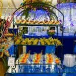 A decorative buffet table by a renowned Catering Company from Baton Rouge features a variety of appetizers. The top and middle tiers of the metal rack display rolled wraps with different fillings. The bottom tier has small cups with skewered fruit. Colorful floral arrangements and illuminated columns are in the background.