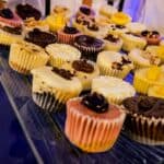 A glass tray from 360 Catering Company holds an assortment of frosted cupcakes in various flavors and colors. The cupcakes, decorated with chocolate, yellow, and white toppings, are arranged in neat rows on the tray—a true testament to the finesse of Baton Rouge caterers.