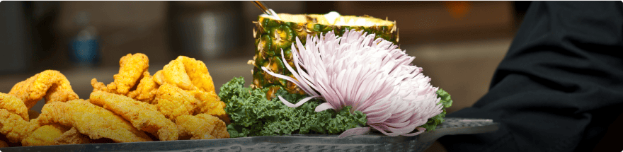 A close-up of a tray featuring deep-fried food items on the left, a whole pineapple used as a bowl with a white chrysanthemum flower next to it in the middle, and leafy greens on the right. The tray, artfully prepared by our catering company, is being carried by a person wearing a black garment.