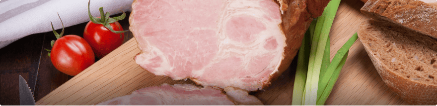 A close-up of a wooden cutting board displays slices of roast pork, two whole tomatoes on the vine, several green onions, and pieces of brown bread. A partially visible striped cloth is situated in the top left corner, showcasing the meticulous presentation by 360 Catering Company from Baton Rouge.