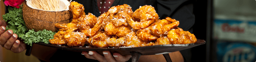 A platter of golden brown fried shrimp, garnished with a sprinkle of shredded coconut and accompanied by a small coconut shell filled with toothpicks. Presented by Baton Rouge Caterers, the platter is held by a person dressed in black.