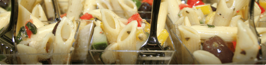 Close-up of multiple servings of pasta salad catered by a top Catering Company Baton Rouge, featuring penne pasta, diced zucchini, red and yellow bell peppers, black olives, and basil. Each small clear container has a black plastic fork placed inside. Perfect for any event venue near Baton Rouge.