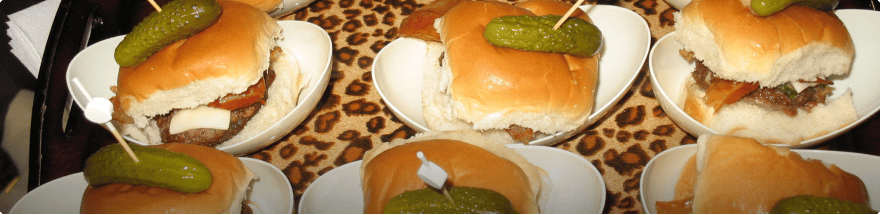 A tray of mini burgers with toppings, including pickles on top, secured by toothpicks. The burgers are placed on small white plates with a leopard print fabric underneath, prepared meticulously by an esteemed Baton Rouge caterer.
