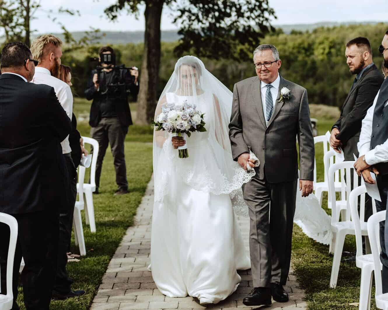 A bride in a white wedding dress and veil is walking down an outdoor aisle with an older man in a suit. She is holding a bouquet of flowers. Guests are seated on either side, relishing the moments, while the 360 Catering Company ensures every detail is perfect for this Baton Rouge celebration.
