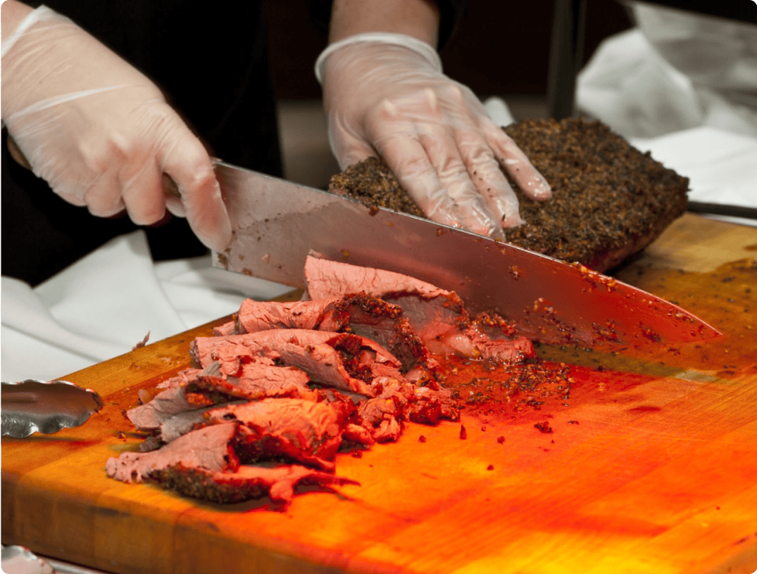 A person wearing gloves is slicing a piece of roast beef on a wooden cutting board with a large knife, expertly showcasing the precision of 360 Catering Company. Several slices of the meat lie arranged in front of the uncut section, with meat juices and seasoning visible on the board.