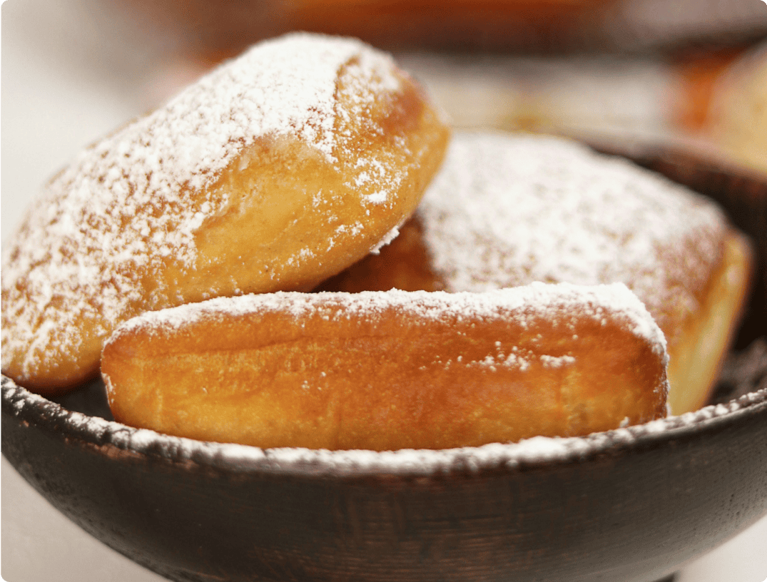 A close-up view of three powdered sugar-covered beignets in a dark wooden bowl. The beignets are golden brown and dusted with a generous amount of powdered sugar, highlighting their texture. Perfect for any catering service or event venue near Baton Rouge, these treats steal the spotlight.