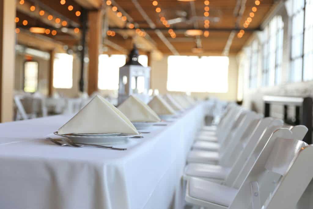 A long dining table is set with a white tablecloth, napkin-folded plates, and silverware. White folding chairs line both sides. A lantern sits in the center of the table. The indoor setting features large windows and warm string lights overhead, expertly arranged by Baton Rouge Caterers.