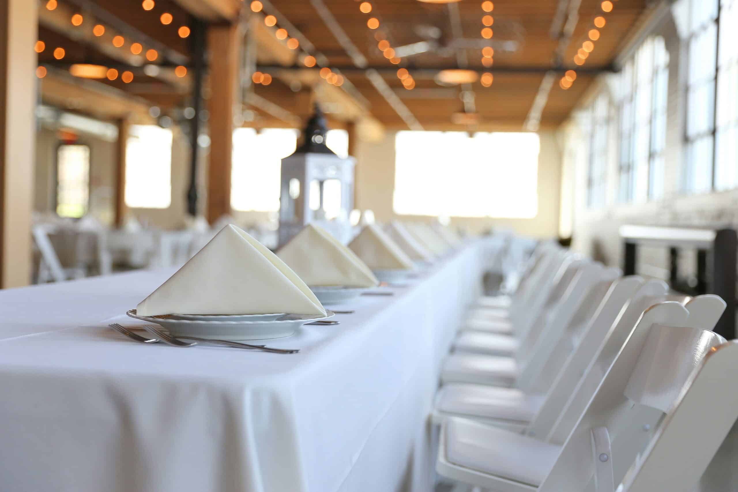 A long dining table is set with a white tablecloth, napkin-folded plates, and silverware. White folding chairs line both sides. A lantern sits in the center of the table. The indoor setting features large windows and warm string lights overhead, expertly arranged by Baton Rouge Caterers.