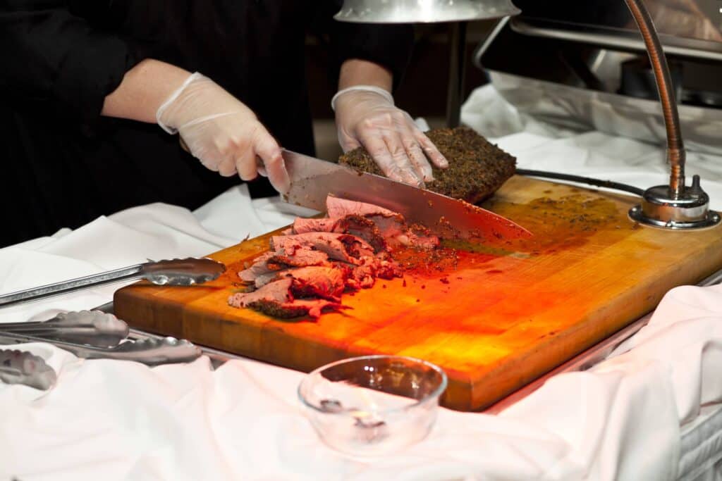 A person wearing gloves from a renowned catering company in Baton Rouge is slicing a piece of seasoned roast beef on a wooden cutting board using a large knife. Tongs and a small, empty glass dish sit beside the board. The area is well-lit, illuminating the food.