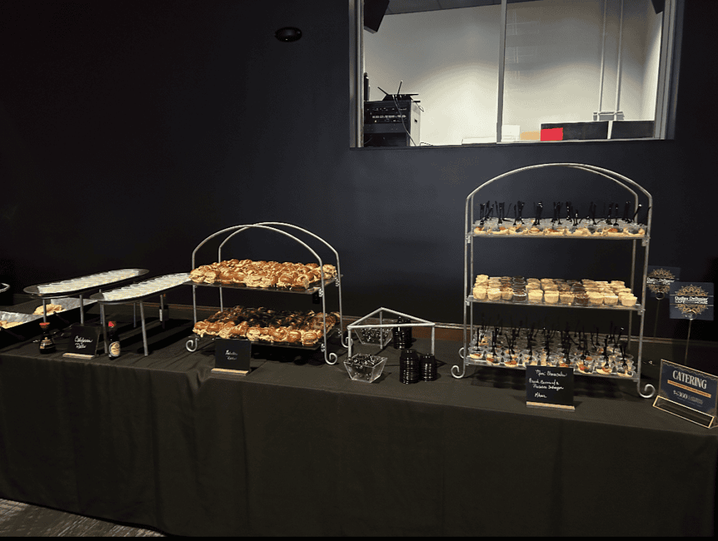 A buffet table is set with an assortment of pastries, desserts, and glasses displayed on two-tier stands and trays. The table is covered with a black tablecloth, and small signs labeling the items are visible, courtesy of Baton Rouge Caterers. The background is a dark-colored wall.