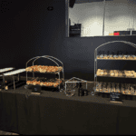 A buffet table is set with an assortment of pastries, desserts, and glasses displayed on two-tier stands and trays. The table is covered with a black tablecloth, and small signs labeling the items are visible, courtesy of Baton Rouge Caterers. The background is a dark-colored wall.