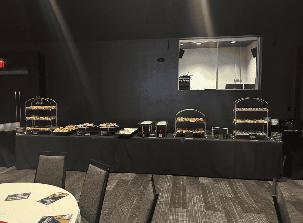 A buffet table with various food items displayed in baskets and trays is set up in a dimly lit room. There are round tables and chairs in the foreground. The walls are dark, and there is a window or opening behind the buffet table, courtesy of Baton Rouge Caterers' exceptional catering service.