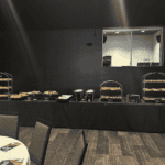 A buffet table with various food items displayed in baskets and trays is set up in a dimly lit room. There are round tables and chairs in the foreground. The walls are dark, and there is a window or opening behind the buffet table, courtesy of Baton Rouge Caterers' exceptional catering service.