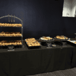 A buffet setup by Baton Rouge Caterers features an array of food items displayed on a long table covered with a black tablecloth. The spread includes trays of pastries, sandwiches, salads, cheese platters, fruits, and other appetizers. A sign on the left reads "Please do not reach over the table.