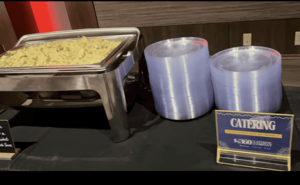 A buffet setup with a metal tray of food, possibly pasta, placed on a black tablecloth. Two large rolls of plastic wrap are next to the tray. A sign on the right reads "Catering" with illegible text below, hinting at top Baton Rouge Caterers providing exceptional Catering Service for your event.