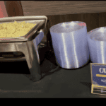 A buffet setup with a metal tray of food, possibly pasta, placed on a black tablecloth. Two large rolls of plastic wrap are next to the tray. A sign on the right reads "Catering" with illegible text below, hinting at top Baton Rouge Caterers providing exceptional Catering Service for your event.