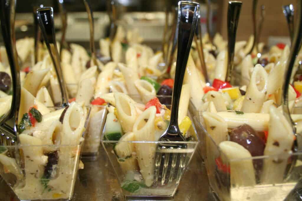 Close-up of several small clear plastic cups filled with pasta salad, consisting of penne pasta, diced red and yellow bell peppers, black olives, and green vegetables. Each cup has a small metal fork standing upright in the salad.