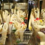 Close-up of several small clear plastic cups filled with pasta salad, consisting of penne pasta, diced red and yellow bell peppers, black olives, and green vegetables. Each cup has a small metal fork standing upright in the salad.