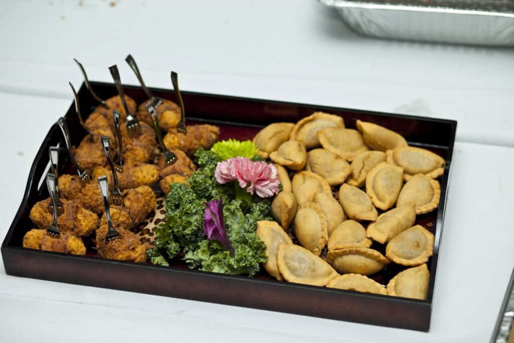 A rectangular serving tray is filled with two types of appetizers. On the left side, there are breaded bite-sized pieces with skewers. On the right side, there are small, half-moon shaped pastries. The tray is garnished with green leafy vegetables and a pink flower.