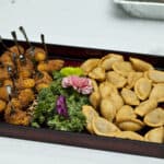 A rectangular serving tray is filled with two types of appetizers. On the left side, there are breaded bite-sized pieces with skewers. On the right side, there are small, half-moon shaped pastries. The tray is garnished with green leafy vegetables and a pink flower.