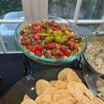 A glass bowl of layered dip topped with cherry tomatoes, olives, and peppers sits behind a plate of tortilla chips on a table.