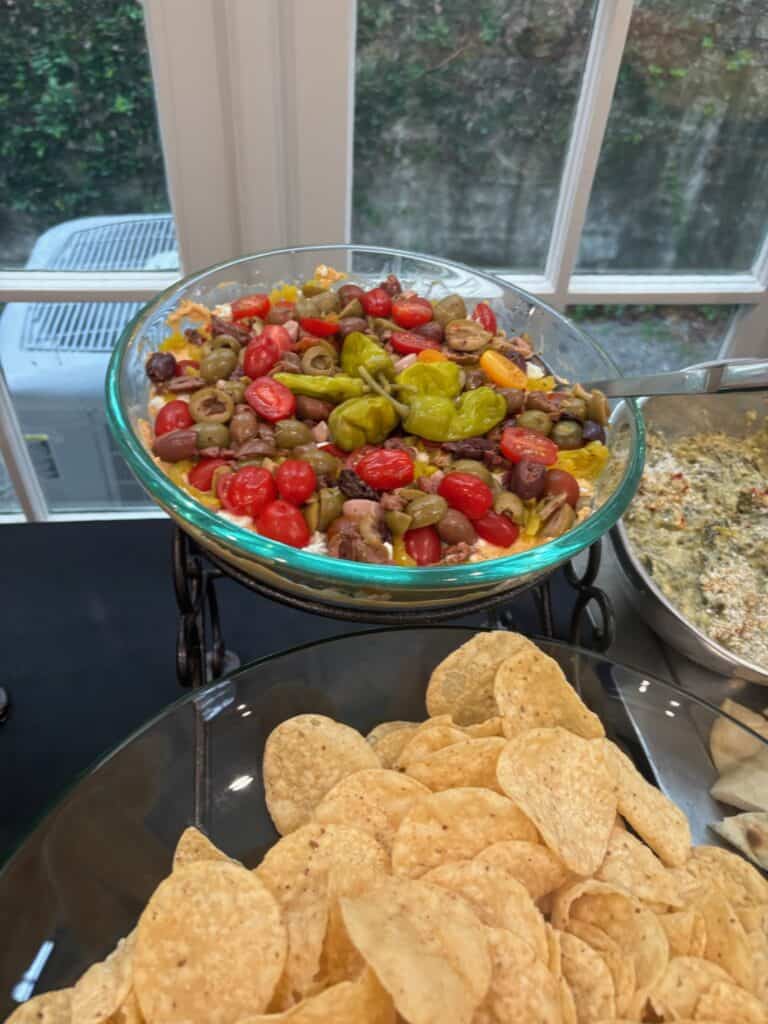 A glass bowl of layered dip topped with cherry tomatoes, olives, and peppers sits behind a plate of tortilla chips on a table.