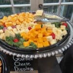 A platter of assorted cheese cubes with greens and grape tomatoes, served with tongs, labeled "Gourmet Cheese Display.