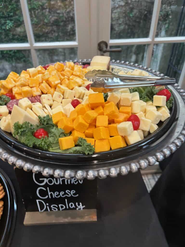 A platter of assorted cheese cubes with greens and grape tomatoes, served with tongs, labeled "Gourmet Cheese Display.