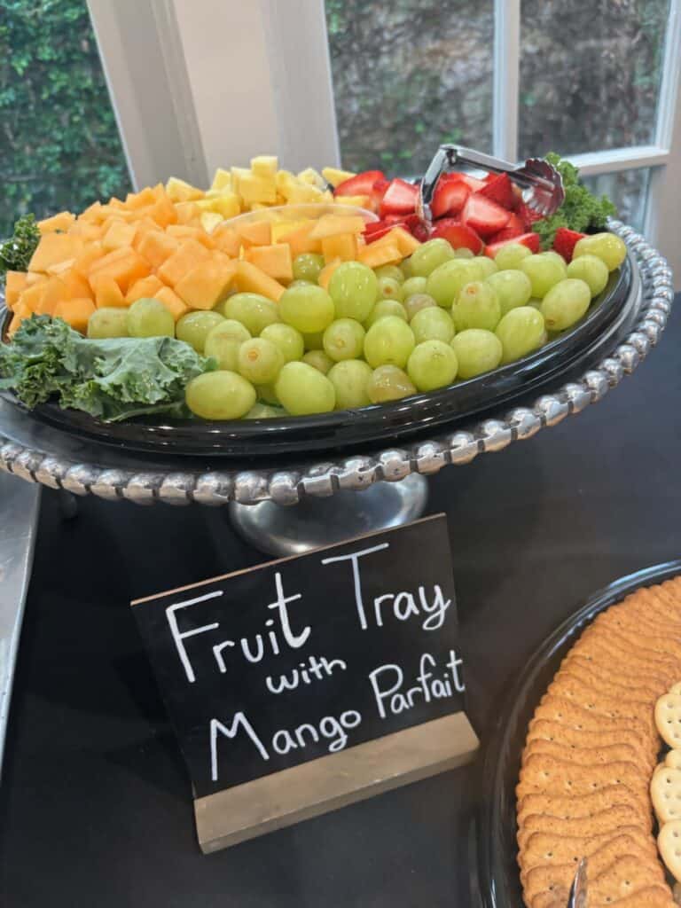 A platter with green grapes, cantaloupe, pineapple, and strawberries, next to a sign that reads "Fruit Tray with Mango Parfait.