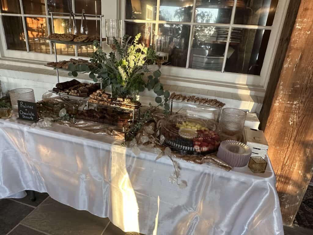A table covered with a white tablecloth displays assorted desserts, fruit, greenery, and stacked trays near a window and wooden beam.
