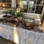 A table covered with a white tablecloth displays assorted desserts, fruit, greenery, and stacked trays near a window and wooden beam.