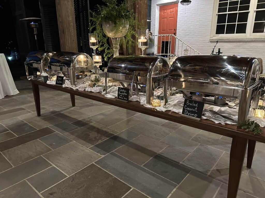 A buffet table with four covered chafing dishes and small chalkboard signs on a stone floor, set up for an event at night.