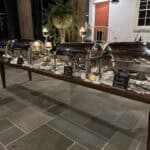 A buffet table with four covered chafing dishes and small chalkboard signs on a stone floor, set up for an event at night.