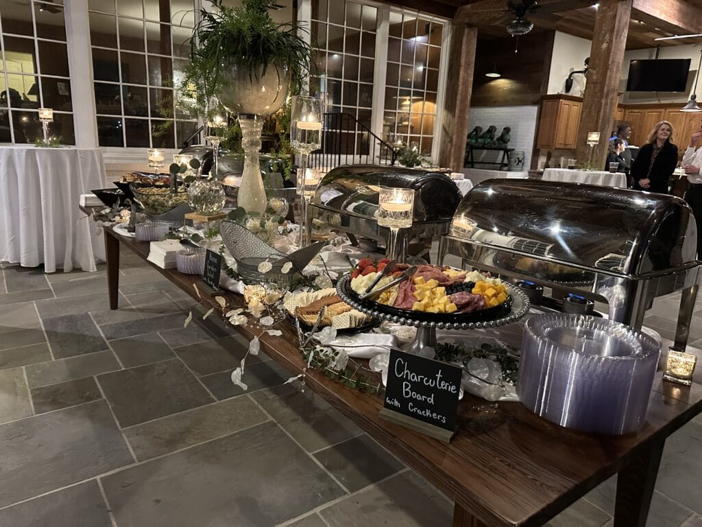 A buffet table with chafing dishes, a charcuterie board, plates, candles, and decorations in a large, well-lit room.