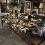 A buffet table with chafing dishes, a charcuterie board, plates, candles, and decorations in a large, well-lit room.