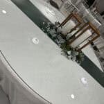 An oval table with a white cloth, a glass top, candles in wooden holders, greenery, and decorative gems as a centerpiece.