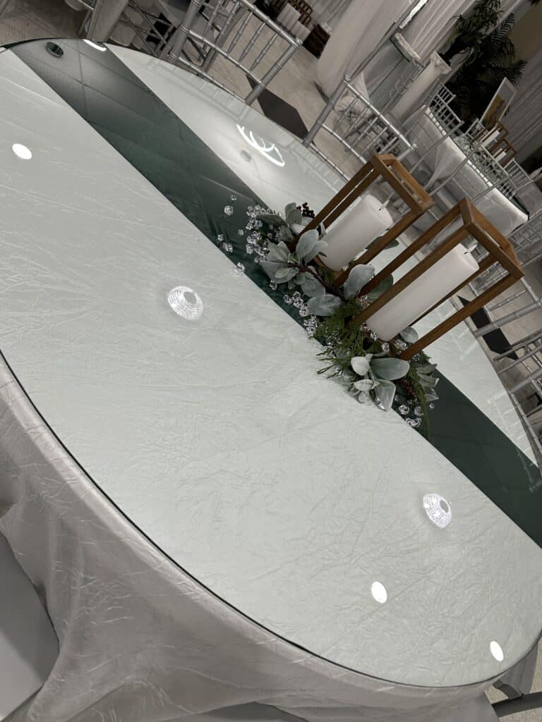 An oval table with a white cloth, a glass top, candles in wooden holders, greenery, and decorative gems as a centerpiece.