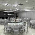 A banquet hall with round tables covered in white linens, silver chairs, and simple centerpiece decorations, under white draped ceiling fabric.