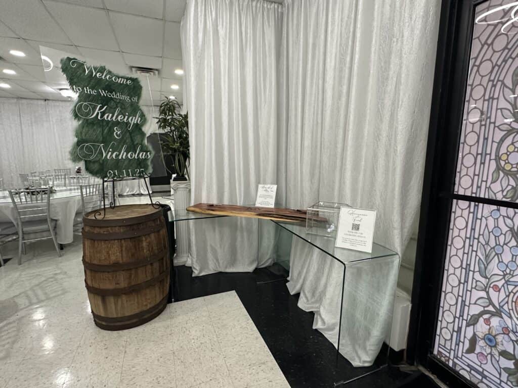 Glass table with guestbook and card box next to a decorative barrel and wedding welcome sign for Raleigh and Nicholas. White curtains in background.