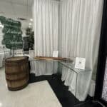 Glass table with guestbook and card box next to a decorative barrel and wedding welcome sign for Raleigh and Nicholas. White curtains in background.