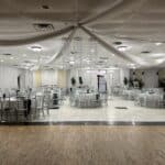 A banquet hall with round tables, silver chairs, white drapes, and ceiling decorations, ready for an event.