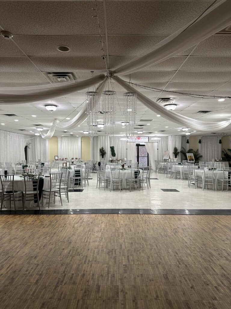 A banquet hall with round tables, silver chairs, white drapes, and ceiling decorations, ready for an event.